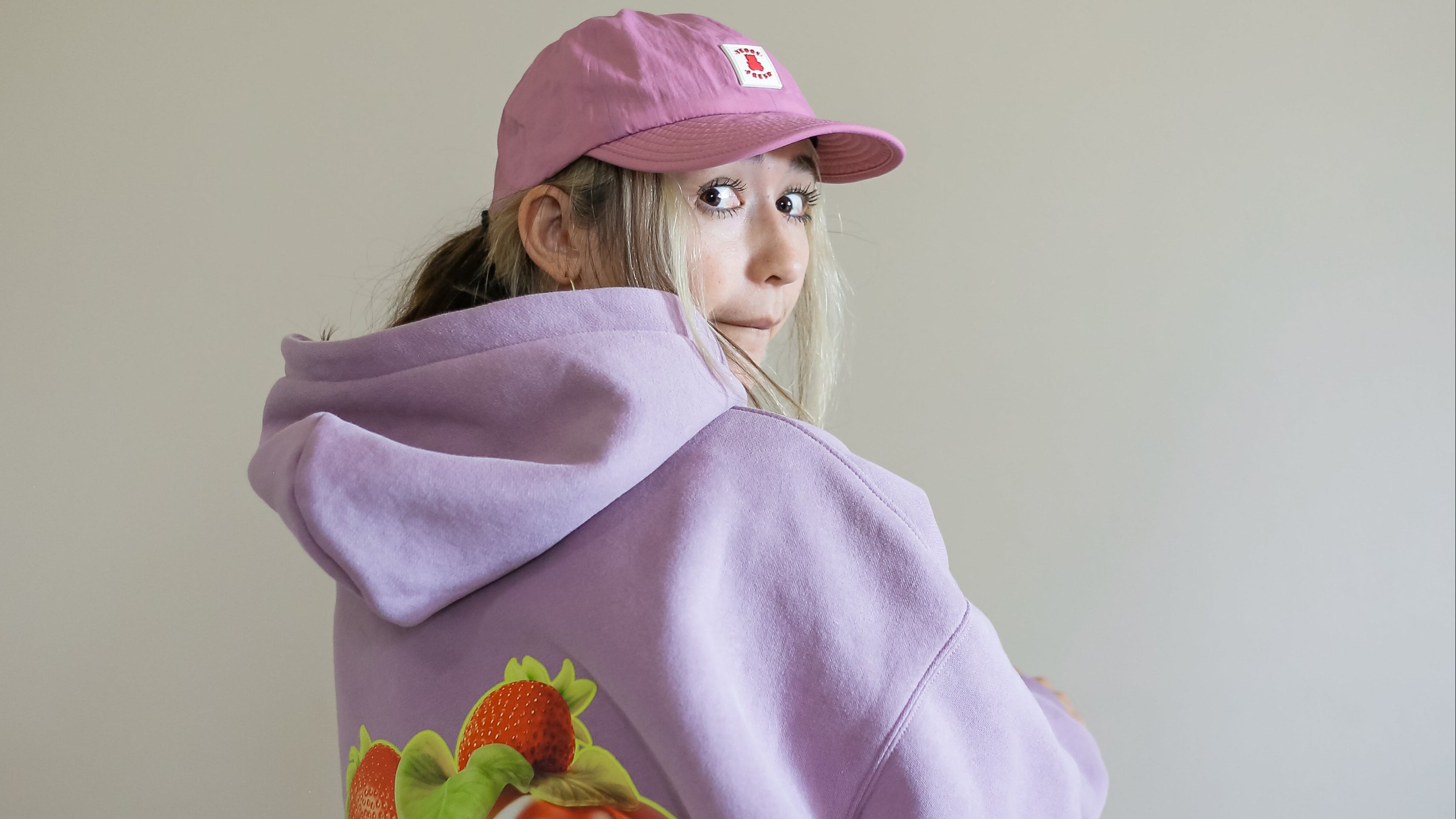 Person wearing a purple hoodie with fruit design and pink cap on a plain background